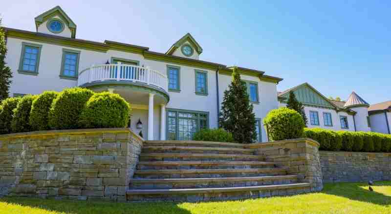 A stone retaining wall that’s lined with neatly trimmed bushes with a large home in the background