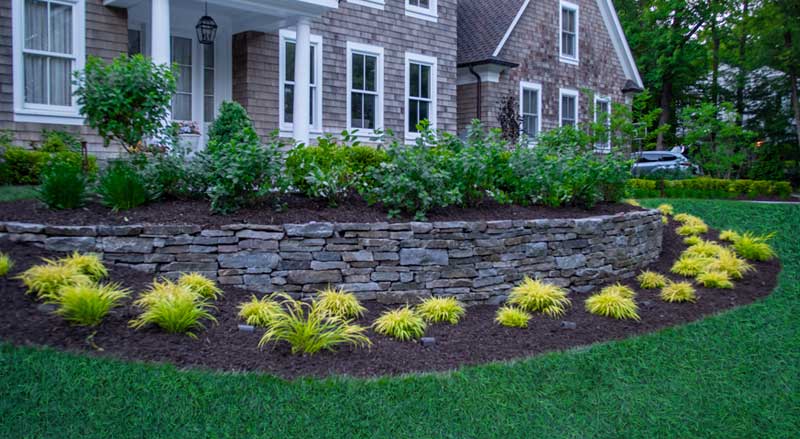 Stone retaining wall in the front yard of a traditional home, accented with lush ornamental grasses for a polished landscape design