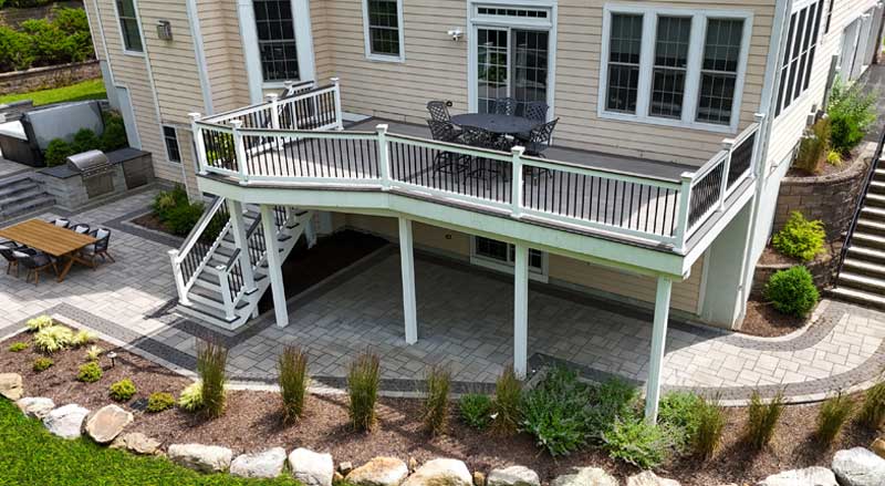 Elevated composite deck connected to a spacious stone patio with dining areas and landscaped pathways