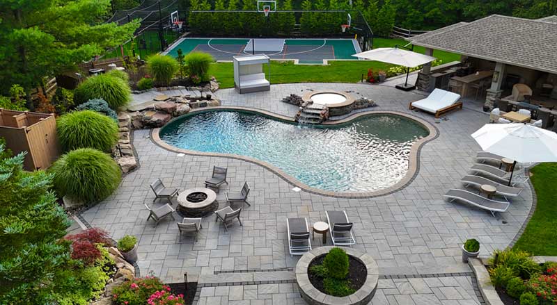 Luxury backyard with freeform pool, fire pit seating, lounge chairs, and adjacent basketball court and pavilion