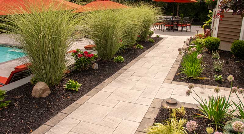Stylish stone walkway bordered by tall ornamental grasses, perennial flowers, and modern patio features