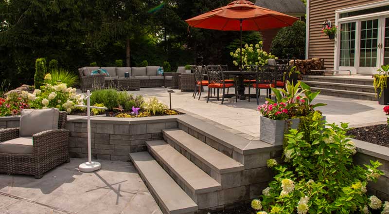 Elevated patio design with tiered steps, wicker lounge furniture, dining area with orange umbrella, and vibrant flower beds