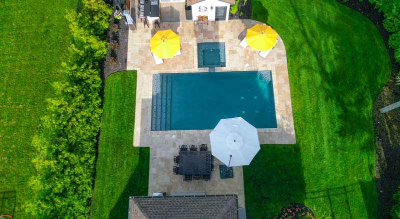 Aerial view of a backyard pool with patio seating and umbrellas, surrounded by lush green lawn