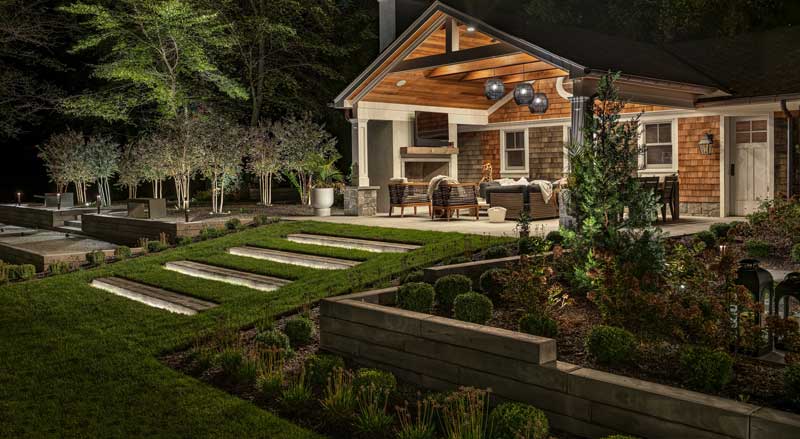 Elegant outdoor living area illuminated at night with landscape lighting, lush gardens, and cozy fall seating under a covered patio