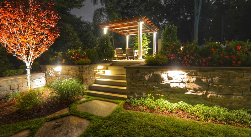 Backyard pergola at night illuminated with warm landscape lighting, surrounded by fall foliage and stone retaining walls