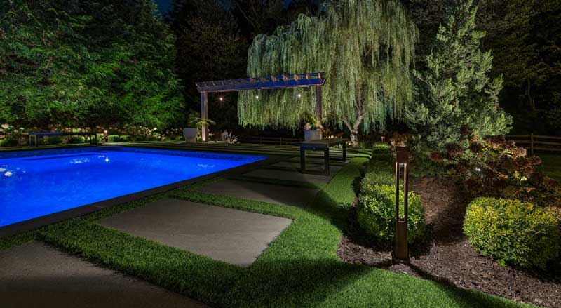 Illuminated backyard pool with modern landscape lighting and lush greenery, showcasing energy-efficient design that adds property value