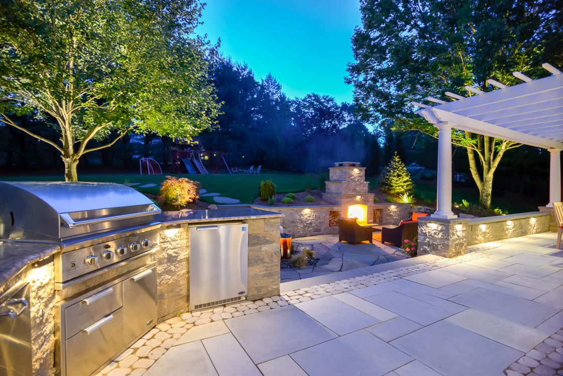 Stone outdoor fireplace and dining room under a pergola with landscape lighting