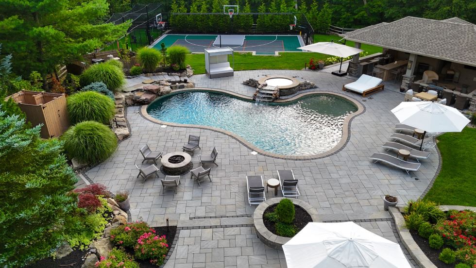 Aerial view of a custom pool landscape with spa, fire pit, outdoor pavilion, patio, plantings, and basketball court
