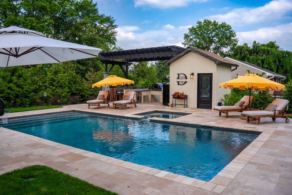 Backyard pool with pool house, outdoor kitchen, pergola, lounge chairs, umbrellas, and stone patio